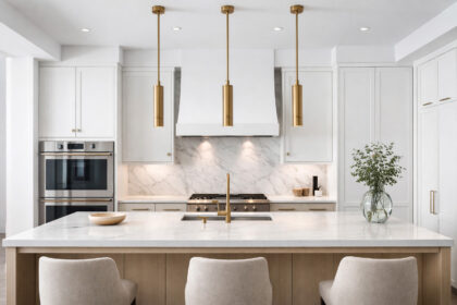 Symmetrical luxury modern kitchen with a white quartz island and three slim brushed brass cylinder pendant lights hanging evenly above in bright natural light