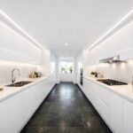 Narrow galley kitchen lighting with white cabinetry and dark stone floor, lit by continuous LED strip lighting on both sides to remove shadows and make the space feel wider and brighter.